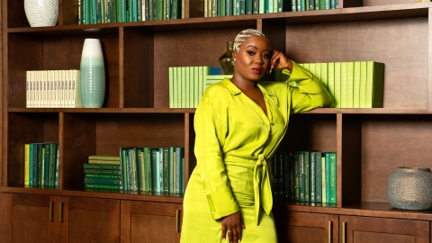 Nadia Sasso, wearing a bright green jumpsuit, poses in front of a bookcase with different shades of green books.