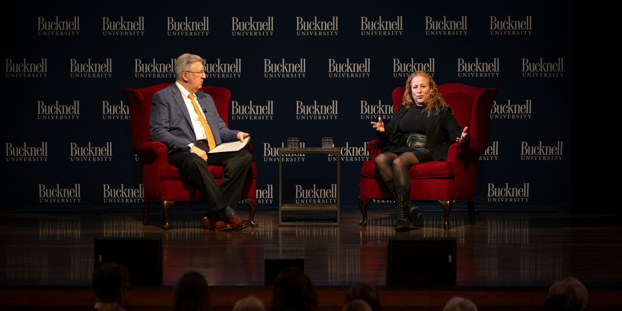 President John Bravman is dressed in a suit and Jodi Picoult is dressed in black and is sitting on red chairs on stage in the Weis Center with a Bucknell University logo backdrop