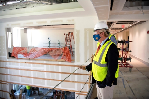 Wearing a hard hat, mask and high-visibility vest, Steve Holmes looks over a balcony at the under-construction Holmes Hall. 