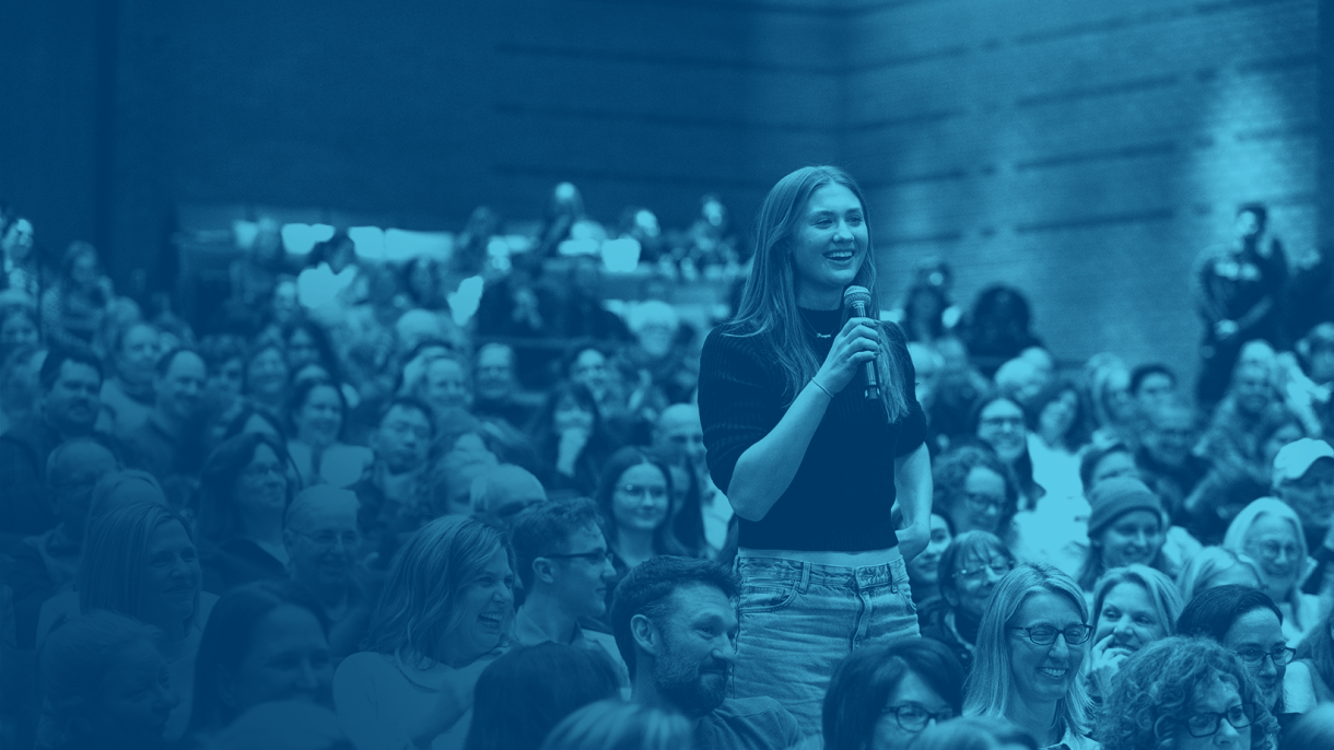 A female student holds a microphone, asking a question to a speaker at a Bucknell Forum event. She is surrounded by a large crowd watching the event in the Weis Center for the Performing Arts.