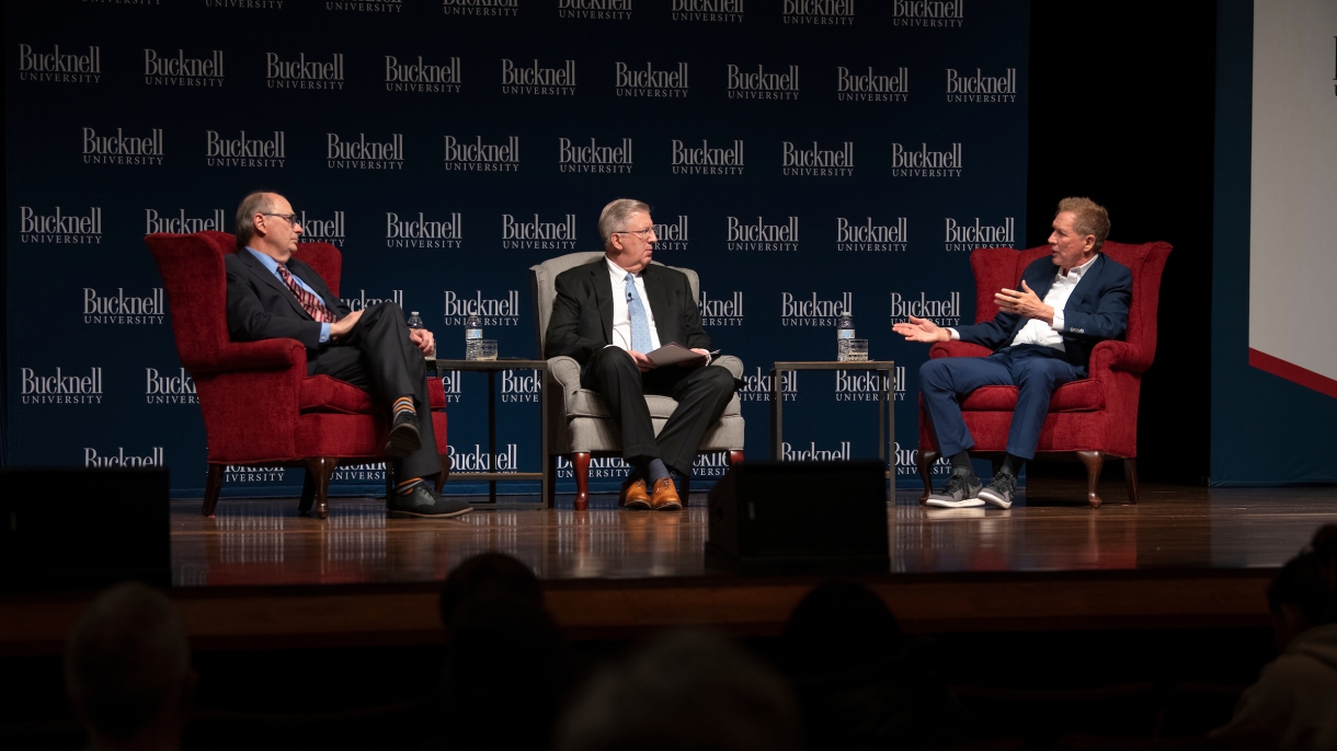 President John Bravman sits on stage at the Weis Center for the Performing Arts between David Axelrod on the left and John Kasich on the right. Kasich is moving his had as if making a point while speaking.