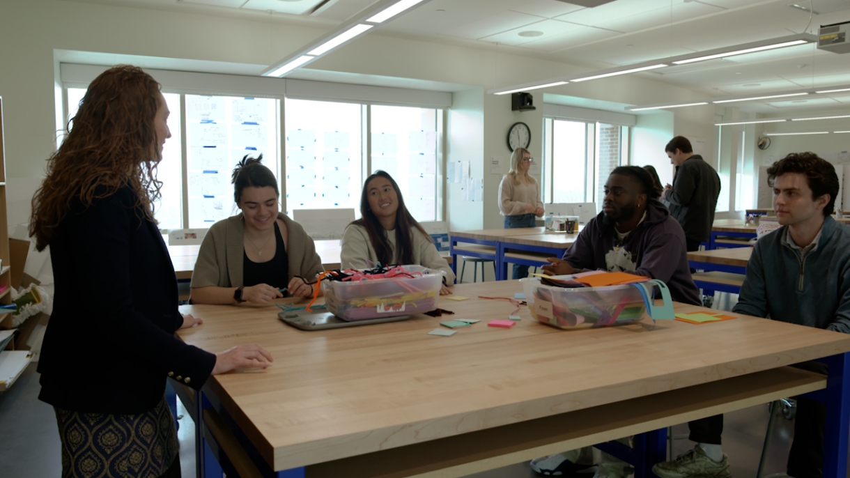 Professor Annetta Grant stands speaking to a group of four students sitting around a high-top table in the Markets, Innovation and Design lab in Holmes Hall