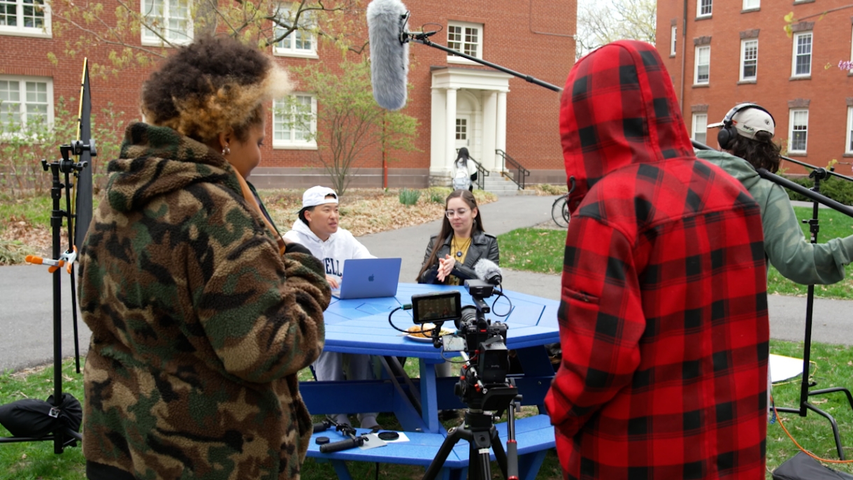 A film crew stands in front of two students sitting at a picnic table outside on the Bucknell campus