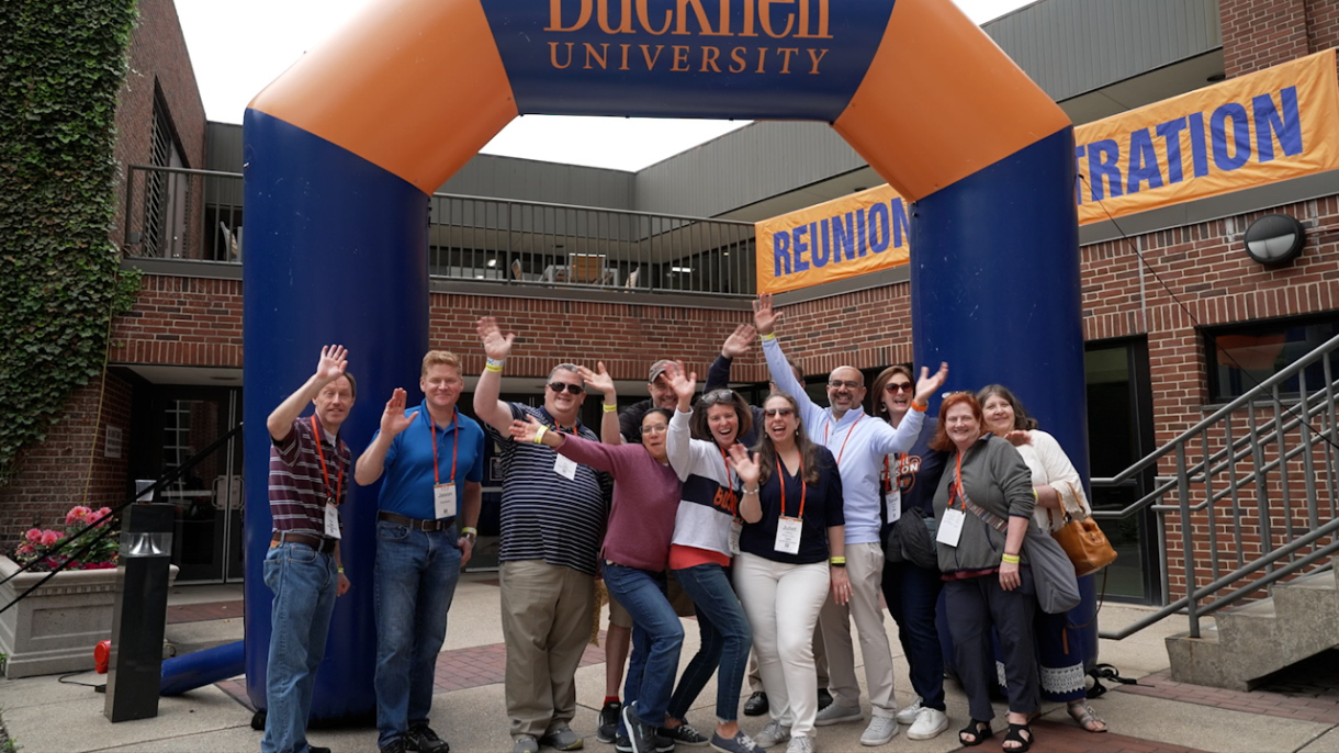 Alumni stand and wave to camera under a blue and orange inflatable arch that says Bucknell University 