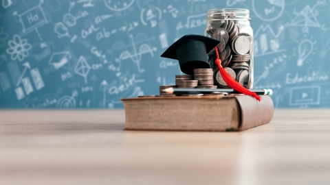 Money in a jar and a tiny graduation cap on top of a book on a table against a board with scientific equations written on it