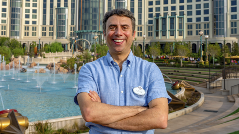 A man in a blue shirt stands with his arms crossed with a blue pool in the background.