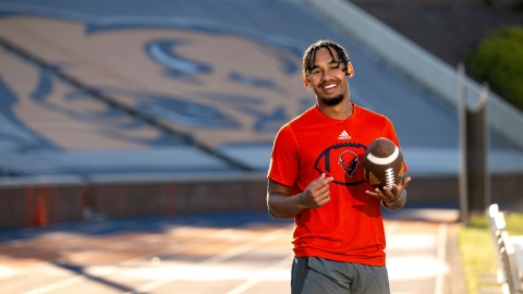 Michael Hardyway &#039;25 wears an orange Bucknell shirt and holds a football as he smiles in Christy Mathewson Memorial Stadium.