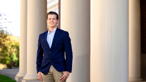 Nick Sperduto &#039;24 wears a navy blazer and tan pants and smiles while standing in front of columns on campus.