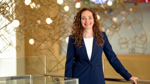 Portrait of a woman wearing a blue suit with her hand on a railing in a bright lobby with a lighted hanging sculpture behind her.