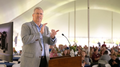 President John Bravman claps his hands and smiles at a podium under a tent at the annual campus picnic.