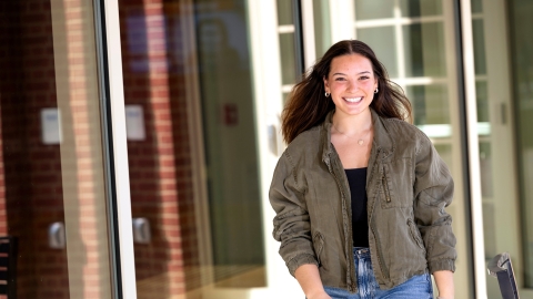 Elizabeth Malley wears a tan jacket and jeans and walks in front of a building on campus with a very big smile.