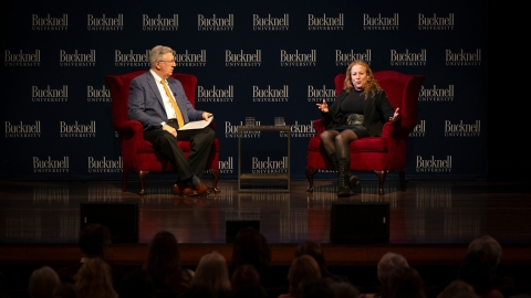 President John Bravman is dressed in a suit and Jodi Picoult is dressed in black and is sitting on red chairs on stage in the Weis Center with a Bucknell University logo backdrop