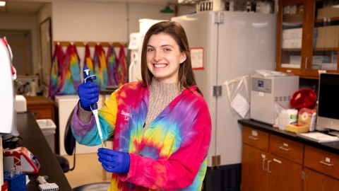 Meghan Catherwood '25 wears a bright, multi-colored tie-dyed lab coat and blue gloves while smiling and holding a pipette in a lab on campus.