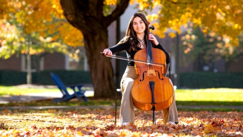 Elle Chrampanis sits on a chair and plays the cello while sitting outside under a tree with golden leaves that hang from the branches and also sit on the ground around her..