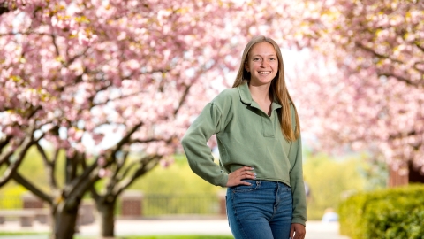 Student Ally Clarke poses to the left-center of the frame standing between two rows of pink cherry blossom trees in full bloom
