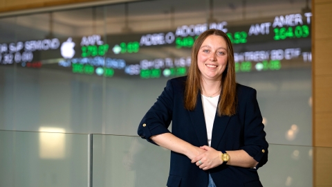 Caitlin Yant '24 stands and smiles in the Moriatry Investment Center in Holmes Hall with a stock ticker behind her.