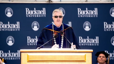 Bucknell president John C. Bravman stands at a podium on the Commencement stage, smiling and wearing sunglasses.