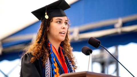Kaia Rendo &#039;23 wears a cap and gown and honor chords as she speaks into a microphone at a podium at Commencement.