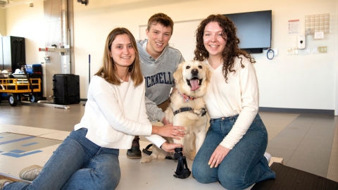 Three students sit and smile with a golden retriever on the ground inside a work space. The dog wears a prosthetic on its front right leg.
