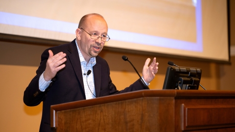 Jon Sheilds stands at a podium with his arms raised as he speaks.