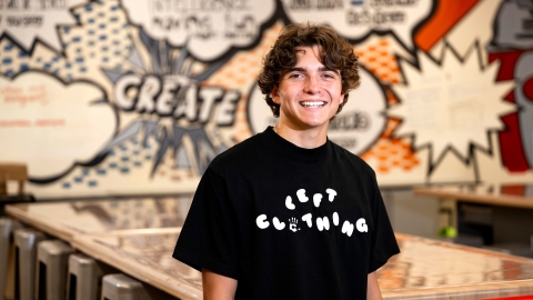 Mikey Brandt wears a black Tshirt with a LEFT Clothing logo printed across his chest. He is in a Makerspace with a graffiti-style wall design behind him.
