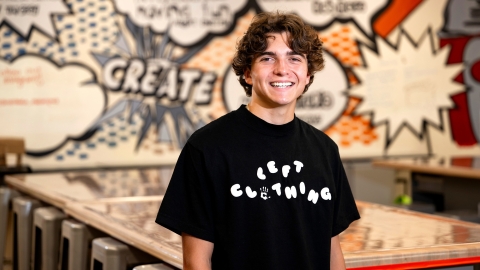 Mikey Brandt wears a black Tshirt with a LEFT Clothing logo printed across his chest. He is in a Makerspace with a graffiti-style wall design behind him.