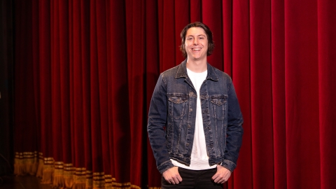 Reid Fournier stands on a stage, a red stage curtain is in the background.