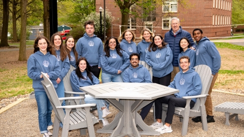 President John Bravman stands with students dressed in matching blue hoody sweatshirts, who are sitting at a table together in Bucknell&#039;s Grove.