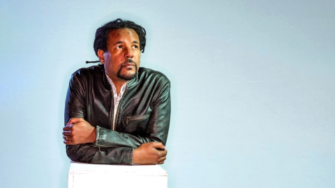 Portrait of author Colson Whitehead leaning forward against a white box, staring upward and off camera to the right.