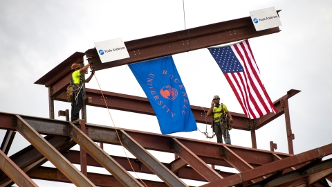 Two construction workers guide a beam into place on top of the building. American and Bucknell University flags hang from the beam.
