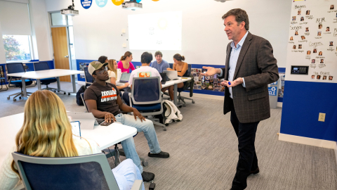 A professor in a suit jacket stands in front of a white board with students surrounding him in a classroom.