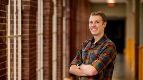 Chris Gregory wears a plaid shirt and stands in a hallway with window and brick trim.