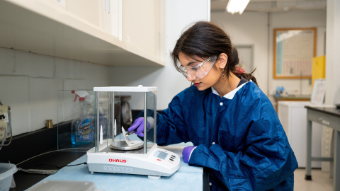 Priyanka Poplai '29 wears a blue lab coat, blue gloves, and safety goggles as she works in a lab space.