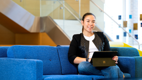 Sophia Bardunias sits on a blue couch in Holmes Hall with a laptop on her lap.