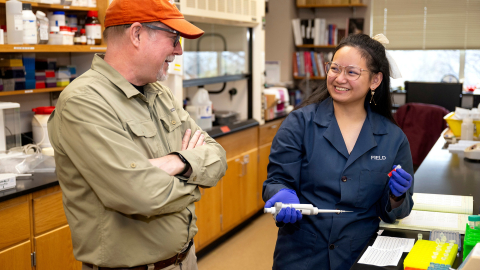 Angel Eunisse Yanga is smiling in a campus lab space wearing a blue lab coat while talking with Professor Ken Field.