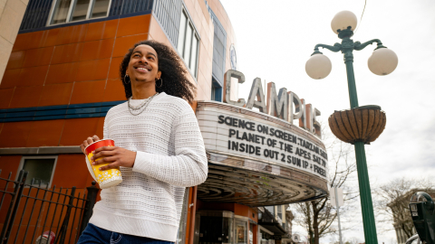 Caleb Warren smiles as he holds a small container of movie theatre popcorn; the marquee of the Campus Theatre is behind him.