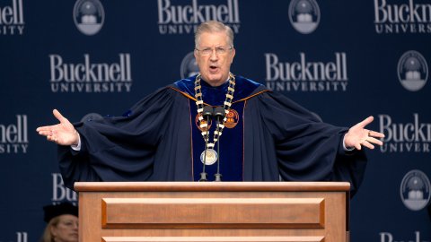 John Bravman speaks at a podium with his arms spread wide. 