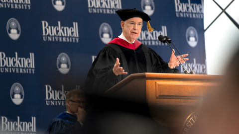 Ken Freeman wears black graduation robes while speaking at a podium.