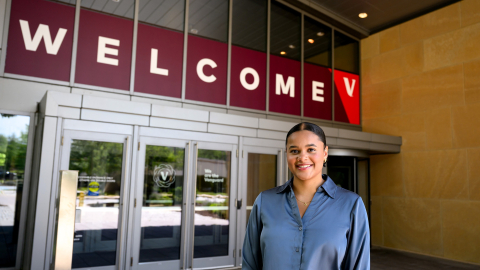 Haden Williams wears a blue shirt and smiles while standing in front of a building that says "Welcome" over glass doors.