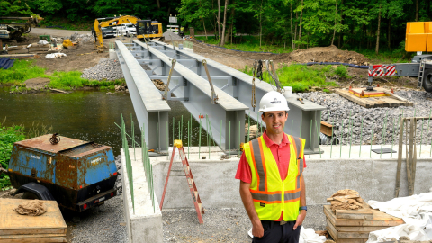 Dominic Viozzi ’27 wears a hard hat and yellow construction safety vest while standing at a bridge construction site.