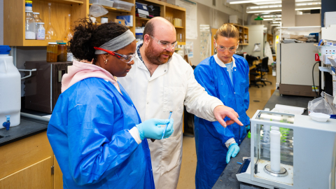 Two students wearing blue lab coats and a professor in a white lab coat stand inside of a laboratory.