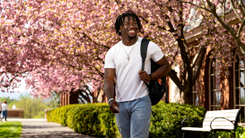 Momo Peters wears a white T-shirt and jeans with a backpack slung over his left shoulder as he smiles and walks on campus with pink cherry blossoms behind him.