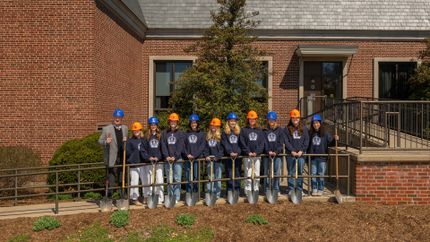a group of students wearing orange and blue hard hats and blue hooded sweatshirts stand in a line together with President John Bravman outside the ELC.