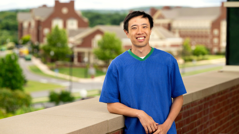 Sing Chun Lee wears a bright blue shirt and leans against a brick wall while smiling on campus