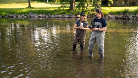 Two researchers wearing chest waders stand in a shallow waterway, one holding a sample bottle and the other checking a handheld device.
