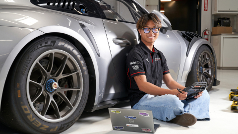 Zach Ebiane sits on the ground with a laptop while leaning against a silver Porsche.