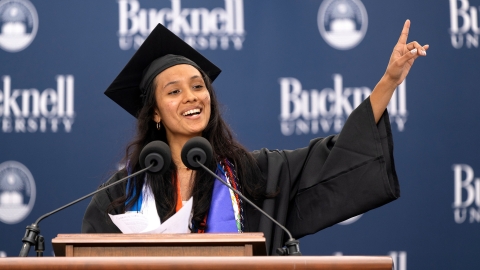 Commencement speaker Gabby Diaz points up with her left hand as she delivers her Commencement speech, wearing a cap and gown