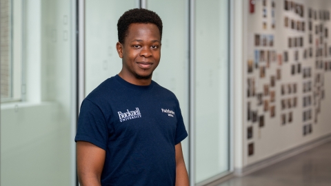 Emanneul Mudungwe stands and smiles in a hallway in Holmes Hall while wearing a navy Bucknell T-shirt