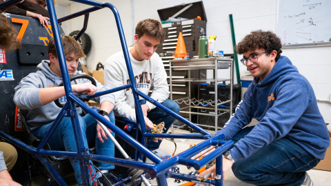 A group of students gather around the frame of a stripped down vehicle.