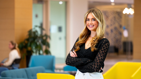 Laura Posh wears a black shirt and white pants and stands with her arms across her torso and smiles; yellow and teal chairs are in the room in the background.